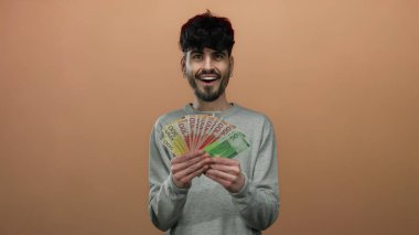 Elinde Norveç banknotları olan gri kazaklı bir adam kahverengi arka plana karşı heyecanlı bir şekilde izole edilmiş, yakışıklı bir ifadeyle para sergiliyor.