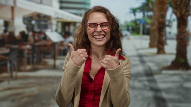 Canlı bir şehir terasında gülümseyen bir kadın palmiye ağaçlarıyla dolu bir restoranda mutlu bir şekilde el kol hareketi yaparak şehir sokaklarında genç ve neşeli bir anı yakalıyor..