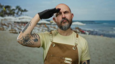 Güneşli bir günde kumlu sahilde dalgalar arkadan vururken kahverengi tulumlu, sakallı ve dövmeli bir adam selamlıyor.