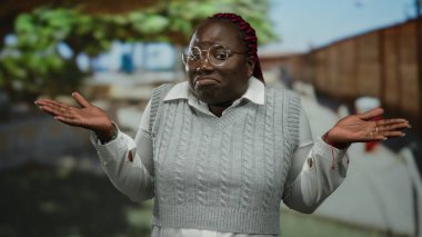 Şehir sokaklarında omuz silken bir kadın, şehir ortamında sorgulayıcı bir ifade sergiliyor, gözlük takıyor ve kırmızı örgülü gri bir kazak giyiyor..