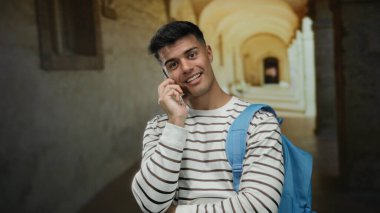 Mavi sırt çantalı genç adam, tarihi üniversite koridorunda telefonla konuşuyor. Rahatlamış atmosferde gülümsüyor ve sohbetin tadını çıkarıyor.