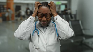 Doktor kadın havaalanı terminalinde gözlük ve steteskop takmış, başı yorgun ve endişeli bir şekilde tek başına dikiliyor.