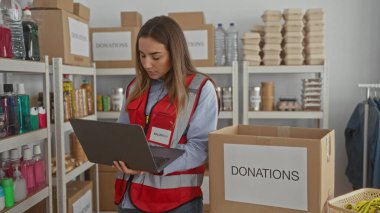 İç mekândaki hayır kurumlarında dizüstü bilgisayar kullanan genç bir kadın. Etrafı kutularla ve malzeme raflarıyla çevrili. Kırmızı yelek giymiş, insani yardım organizasyonları düzenliyor.