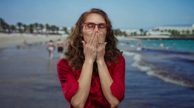 Plajda kırmızı tişörtlü ve gözlüklü bir kadın açık gökyüzünün altında deniz kenarında bir arka planda kameraya öpücük yolluyor..