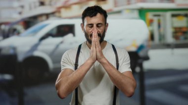 Sakallı İspanyol bir adam canlı bir sokakta namaz kılıyor. Pantolon askısı ve arka planda bulanık yayalar gibi beyaz bir gömlek giyiyor..