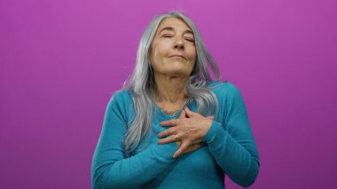 Mavi kazaklı, yaşlı bir kadın. Yalıtılmış pembe arka planda el ele tutuşuyor. Minnettarlığını ve huzurunu ifade ediyor..