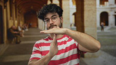 Çizgili tişörtlü sakallı genç adam üniversite bahçesinde kemerli bir jest yapıyor. Akademik ortamda düşünceli bir mola veriyor..
