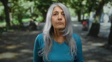 Gri saçlı yaşlı bir kadın parkta duruyor, mavi bir kazak giyiyor, yemyeşil ağaçlarla çevrili, sükunet ve doğa duygusunu taşıyor..