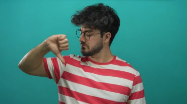 Çizgili tişörtlü, sakallı genç adam yeşil arka plana başparmak işareti yapıyor. Düşünceli bir bakışla karışık tepkiler veriyor..