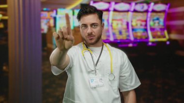 Sakallı ve steteskoplu genç beyaz erkek doktor kumarhanede duruyor. Arka planda kumar makineleri var. Sağlık ve kumar ortamlarının benzersiz bir zıtlığını oluşturuyor..