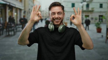 Sakallı ve kulaklıklı genç adam şehrin arka planında bulanık bir jest yaparken gülümsüyor. Açık havada tasasız ve rahat bir atmosfer izlenimi veriyor..