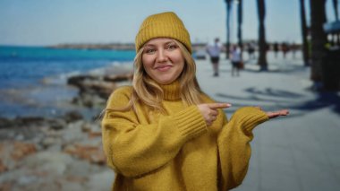 Sarı kazaklı ve bereli kadın deniz kenarındaki gezinti alanında palmiye ağaçları ve okyanusu işaret ediyor. Manzaralı plaj ve canlı açık hava manzarası sergiliyor..