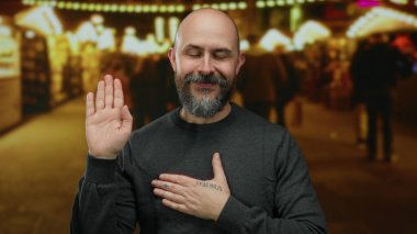 Aydınlatılmış bir şehir sokağında güvenle duran bir adam, el kaldırmış el kol hareketi yapıyor, diğeri göğsünde kel, sakallı, sakin bir ifade takınıyor ve günlük giysiler giyiyor..