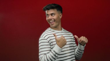 Çizgili bir süveter içinde kırmızı bir arka planda baş parmağıyla gülümseyen genç adam, neşeli bir ifadeye izole edilmiş özgüven ve pozitiflik sergiliyor.