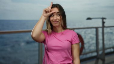 Genç bir kadın deniz manzaralı bir gezide ezik bir jest yapıyor, pembe bir gömlek giyiyor, kendine güven ve mizah yayıyor, su ve açık hava manzarası ile çevrili..