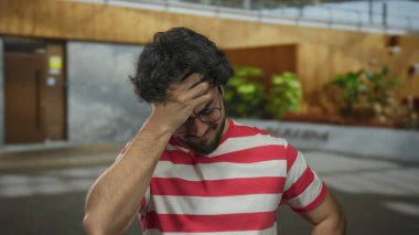 Güneşli bir günde açık havada çizgili tişörtlü, sakallı genç bir İspanyol, başını bitki ve cam yansımalarla dolu modern bir şehir ortamında tutarken stresli görünüyor..