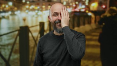 Sakallı bir adam gece şehir sokağında gülüyor. Arka planda bulanık ışıklar var. Canlı ve neşeli bir şehir manzarası yaratıyor..
