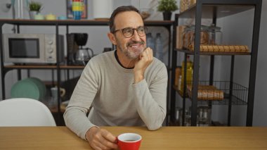 Ofis ortamında, içinde raflar ve modern dekorlarla çevrili bir kahve fincanı olan bir masada oturan olgun İspanyol bir adam rahat ve profesyonel bir atmosfer sunuyor.