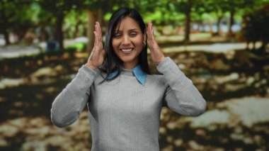 Parkta gülümseyen genç bir kadın, güneşli bir günün tadını çıkarırken elleriyle el kol hareketi yapıyor. Etrafı yemyeşil ağaçlarla çevrili ve doğa mutluluğu ve yaratıcılığı temsil ediyor..