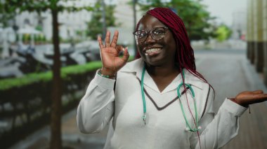 Açık şehir caddesinde saçları örgülü ve gözlüklü bir kadın, steteskop takıyor ve avucunun içini işaret ediyor, canlı bir sokak arkaplanı sergiliyor..