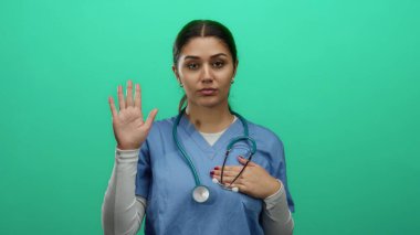 Genç İspanyol kadın hemşire, elini kalbe koyup yemin ediyor ve elini kaldırıyor, yeşil bir zemine dayanıyor, bu da profesyonellik ve kendini adamışlığı gösteriyor..
