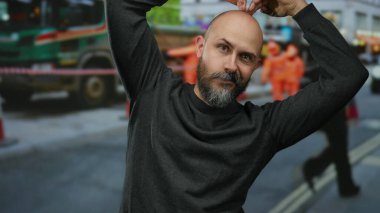 Sakallı kel adam, şehir sokağında güvenle poz veriyor. Araçların geçmişi bulanık ve şehir merkezinde insanlar var..