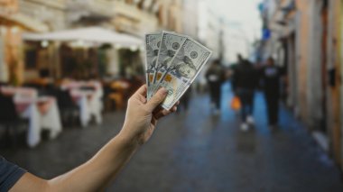 Arka planında restoran terası olan hareketli bir caddede Amerikan banknotlarını tutan adam, sıradan açık hava yemeklerini ve kentsel finansal işlemleri gösteriyor..