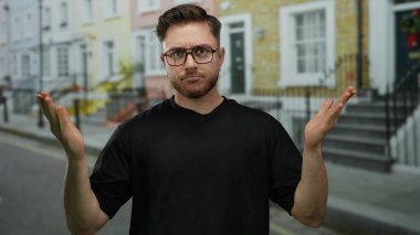 Sakallı ve gözlüklü genç bir adam şehir sokaklarında, açık avuçlarla kafa karışıklığını dile getiriyor..