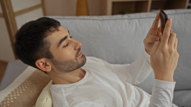 Genç adam evde dinleniyor, rahat oturma odasında akıllı telefon kullanıyor, günlük kıyafetler giyiyor, modern bir yaşam tarzını ve rahat bir ev atmosferini yansıtıyor..