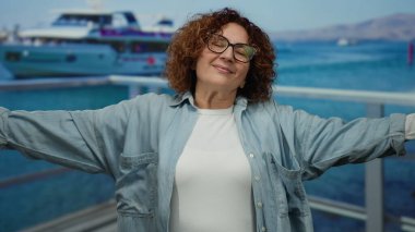 Kadın deniz kenarındaki gezinti güvertesinde kollarını açmış arka planda botlar ve mutluluk ve özgürlüğü yansıtan gözlüklerle okyanus manzarasını seyrediyor..
