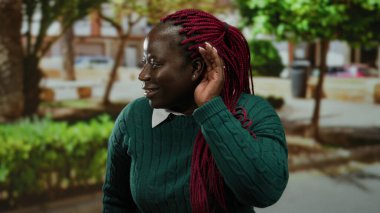 Kulaktan kulağa dikkatle dinleyen, yeşil süveter giyen, ağaçlarla çevrili ve şehir dışında yaşayan, canlı kırmızı örgüler sergileyen bir kadın..