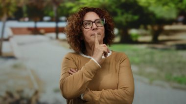 Gözlüklü İspanyol kadının güneşli bir parkta yaratıcı bir jesti var. Açık havada canlı bir yeşilliğin ortasında iç gözlem ve düşüncelilik sergiliyor..