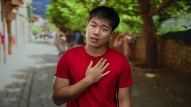 Kırmızı tişörtlü genç adam şehir caddesinde duruyor. Duygularını ifade ediyor. Göğsünde bulanık insanlar ve açık hava ışıkları altında ağaçlar var.