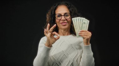 Gözlüklü orta yaşlı bir kadın siyah bir geçmişe karşı iyi bir jest yaparak Amerikan doları tutuyor. Bu da güven ve finansal başarıyı gösteriyor..