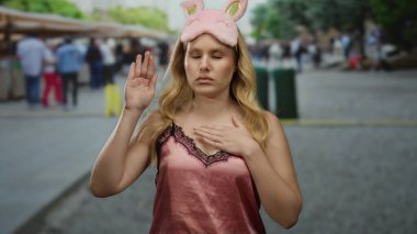 Gecelikli genç sarışın bir kadın işlek bir şehir caddesinde duruyor, pembe bir göz maskesi takıyor ve bulanık şehir kalabalığının arasında rehin jesti yapıyor..