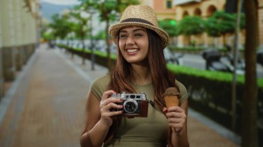 Güneşli bir günde şehir manzarasını keşfederken kamera şapkası ve dondurmayla gülümseyen kadın anılarını yakalıyor..