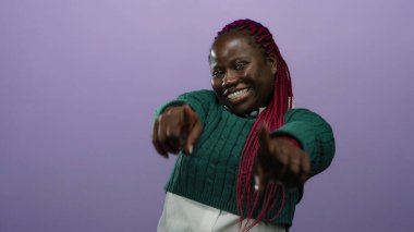 Canlı kırmızı örgüleri ve yeşil süveteri ile mor izole edilmiş arka planda güvenle gülümseyen kadın kameraya neşeli bir şekilde bakıyor..