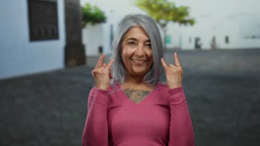 Gri saçlı yaşlı kadın canlı bir sokak ortamında taştan jest yapıyor. Pembe bir kazak giyiyor. Sıradan bir şehir ortamında neşe ve güven saçıyor..