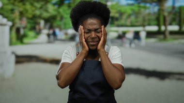 Açık şehir caddesinde elleri açık önlük giyen bir kadın, bulanık arka plan ve yeşil yeşillikler arasında duygu ve içgözlem taşıyor..