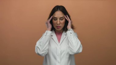Beyaz önlüklü bir kadın, izole edilmiş kahverengi bir arka planda stres yaşıyor. Konsantrasyon anını vurguluyor..
