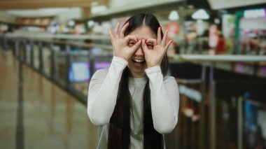 Uzun saçlı, gözlüklü, elleri açık alışveriş merkezinin içinde beyaz gömlekli, hareketli atmosfer ve canlı mağazalarla çevrili bir kadın.