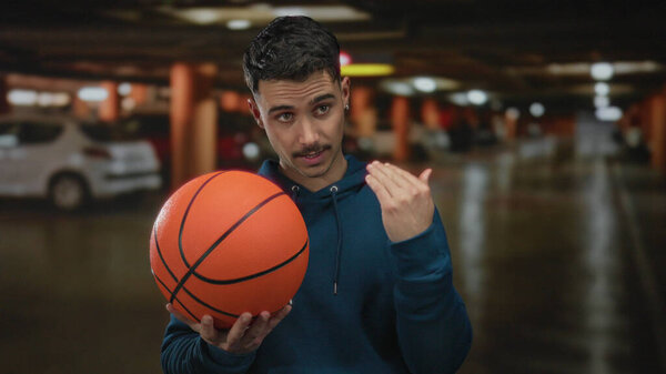 Молодой латиноамериканец держит баскетбольный мяч на крытой парковке, делает жест рукой, как будто зовет кого-то приехать..