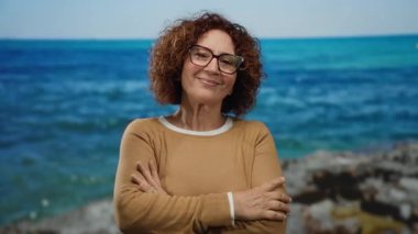 Orta yaşlı İspanyol bir kadın, deniz kenarında, mutlu bir ifadeyle, gözlük takıyor ve kahverengi bir süveter giyiyor, canlı mavi bir deniz ve gökyüzünün zeminine karşı..