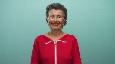 Kıdemli kadın gri saçlı, kırmızı tişörtlü mavi arka planda duran neşeli bir ifade ve pozitif tavır sergileyen biri..