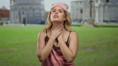 Uyku maskesi takmış bir kadın boğazına dokunuyor güneşli bir günde sidik kulesinin önünde duruyor, rahatsızlık ya da acı veriyor..