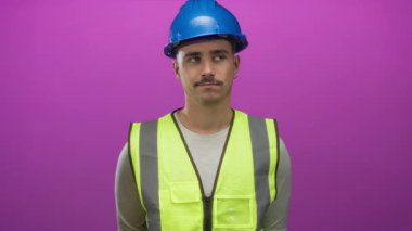 Yansıtıcı yelek ve kasketli genç adam, izole edilmiş pembe arka plan duvarına karşı duruyor, düşünceli ve odaklanmış görünüyor, profesyonel ve kendinden emin bir tavır sergiliyor..