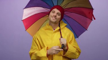 Renkli şemsiyelerin altında gülümseyen genç adam mor arka planda sarı yağmurluk giyiyor..