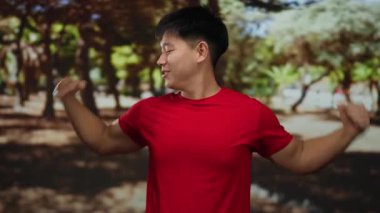 Parkta kaslarını esneten genç Çinli bir adam ağaçlarla çevrili kırmızı gömlekli ve güneş ışığıyla çevrili barışçıl doğal ortamda güç ve kendine güven gösteriyor..