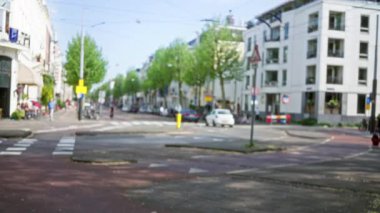 Güneşli gökyüzü altında insanların ve arabaların olduğu bulanık bir Avrupa caddesi, modern binalar ve yemyeşil ağaçlarla hareketli bir şehir manzarası sergiliyor.