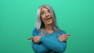 Gri saçlı, mavi kazaklı bir kadın canlı yeşil arka plana karşı yan yan bakıyor. Yön ve pozitifliği neşeli bir ifadeyle aktarıyor..